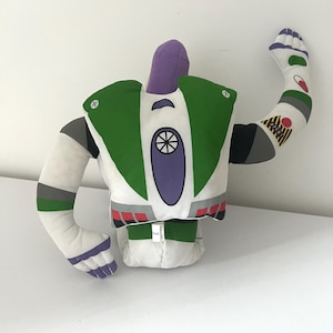May include: A plush Buzz Lightyear toy, featuring a white body with green, purple, and gray accents. The toy has one arm raised, showcasing details like the character's signature design. The toy is a soft, stuffed figure.
