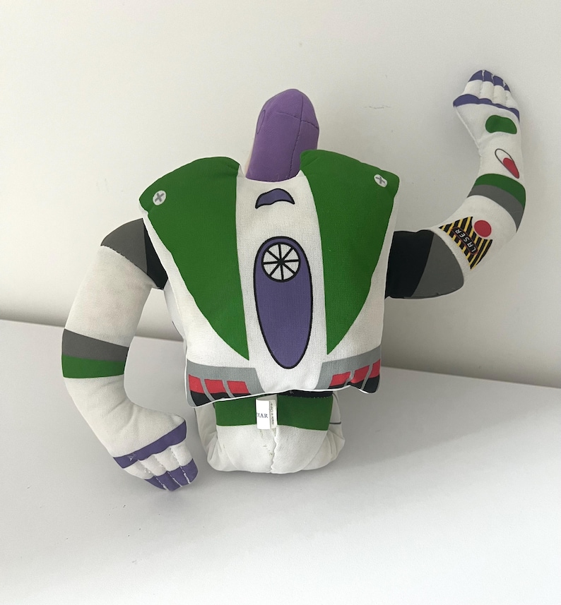 May include: A plush Buzz Lightyear toy, featuring a white body with green, purple, and gray accents. The toy has one arm raised, showcasing details like the character's signature design. The toy is a soft, stuffed figure.