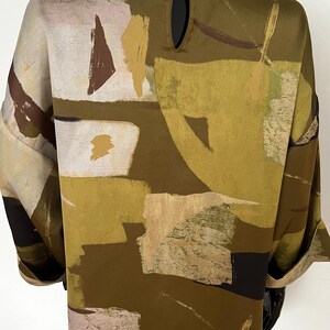 May include: A loose-fitting top with a unique abstract pattern in shades of olive green, brown, beige, and black. The design features geometric shapes and brushstroke-like patterns. The top has a keyhole detail at the back and rolled-up sleeves.