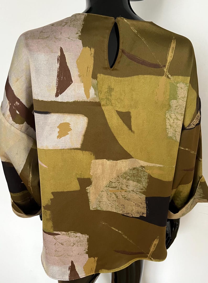 May include: A loose-fitting top with a unique abstract pattern in shades of olive green, brown, beige, and black. The design features geometric shapes and brushstroke-like patterns. The top has a keyhole detail at the back and rolled-up sleeves.