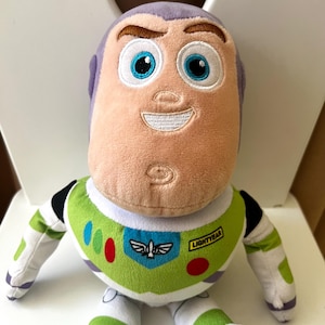 May include: A plush Buzz Lightyear doll, featuring a light peach face, purple head, and a green and white spacesuit. The doll has embroidered facial features, including blue eyes and a smiling mouth. The chest of the spacesuit has a Lightyear logo.