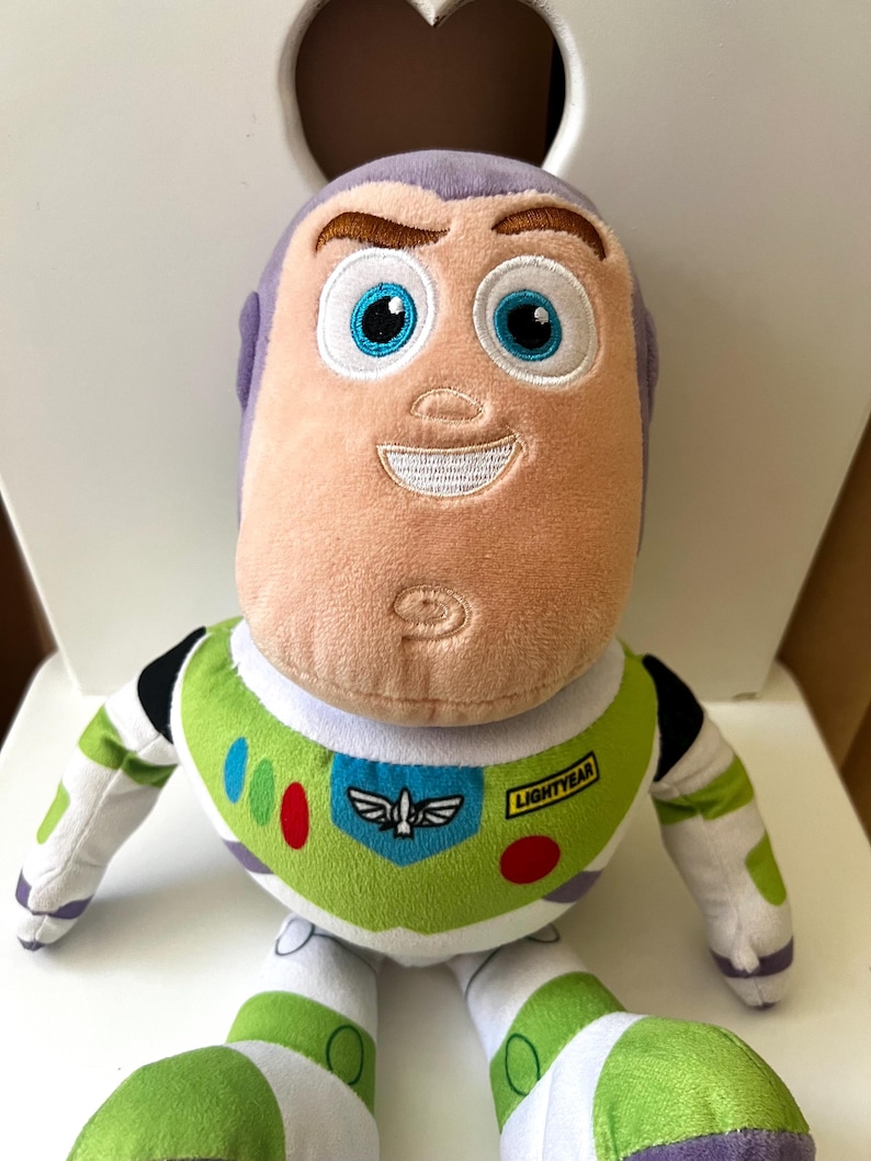 May include: A plush Buzz Lightyear doll, featuring a light peach face, purple head, and a green and white spacesuit. The doll has embroidered facial features, including blue eyes and a smiling mouth. The chest of the spacesuit has a Lightyear logo.