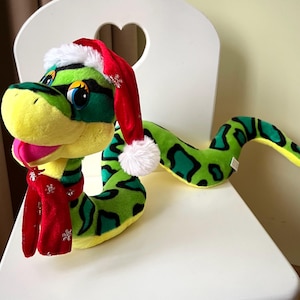 Può includere: Un peluche a forma di serpente con un tema natalizio. Il serpente è principalmente verde con macchie nere e un ventre giallo. Indossa un cappello da Babbo Natale rosso con finiture bianche e una sciarpa rossa abbinata con dettagli a fiocco di neve. Il serpente è posizionato su una sedia bianca.