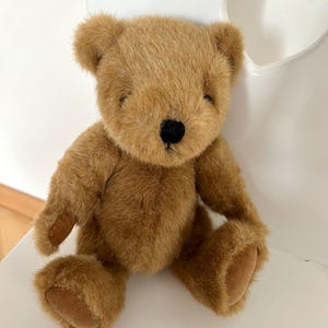 Peut inclure: Un ours en peluche brun avec un nez noir et des yeux fermés. L'ours est fait d'une matière douce et moelleuse. Il est assis sur une surface blanche. Les pattes et les pieds de l'ours sont d'une nuance de brun plus claire.