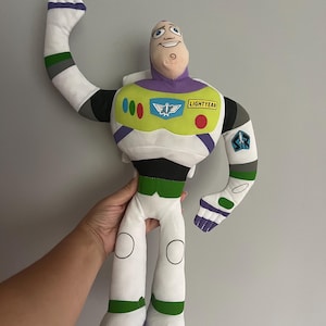 May include: A plush Buzz Lightyear doll, featuring a white and green spacesuit with purple and black accents. The doll has a smiling face and the word "LIGHTYEAR" on the chest. One arm is raised in a waving gesture.