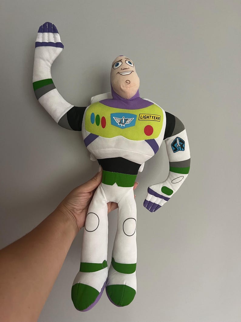 May include: A plush Buzz Lightyear doll, featuring a white and green spacesuit with purple and black accents. The doll has a smiling face and the word "LIGHTYEAR" on the chest. One arm is raised in a waving gesture.