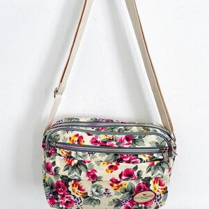 Op de afbeelding: Bloemen crossbodytas met een cr&egrave;mekleurige canvas riem. De tas heeft een beige achtergrond met een patroon van roze, rode en gele bloemen met groene bladeren. Het heeft drie vakken met ritssluiting.