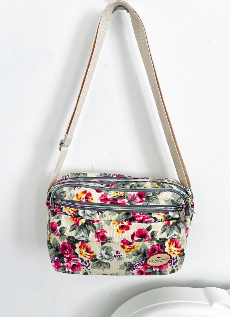 Op de afbeelding: Bloemen crossbodytas met een cr&egrave;mekleurige canvas riem. De tas heeft een beige achtergrond met een patroon van roze, rode en gele bloemen met groene bladeren. Het heeft drie vakken met ritssluiting.