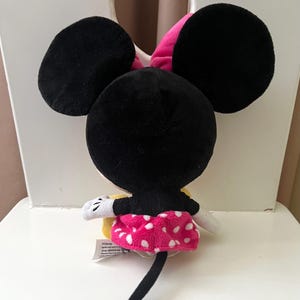 May include: A plush Minnie Mouse toy, viewed from the back, with large black ears and a pink bow. She wears a pink skirt with white polka dots, white gloves, and a black tail. The toy is sitting on a white chair.