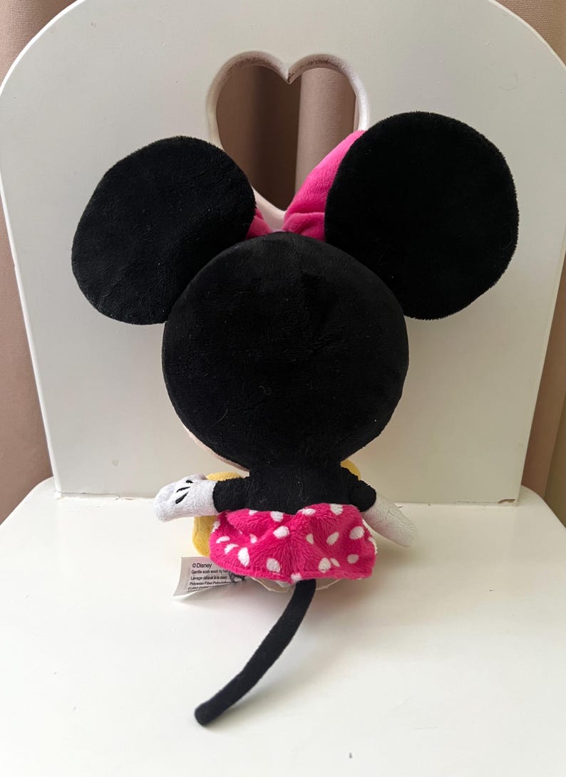 May include: A plush Minnie Mouse toy, viewed from the back, with large black ears and a pink bow. She wears a pink skirt with white polka dots, white gloves, and a black tail. The toy is sitting on a white chair.