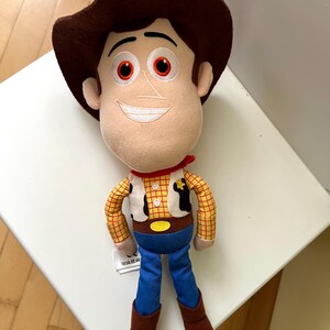 Puede incluir: Un peluche de un personaje de dibujos animados de vaquero. El juguete tiene un sombrero marr&oacute;n, una cara beige con una expresi&oacute;n sonriente y un pa&ntilde;uelo rojo. El personaje viste una camisa a cuadros amarilla, un chaleco blanco, pantalones azules y botas marrones.