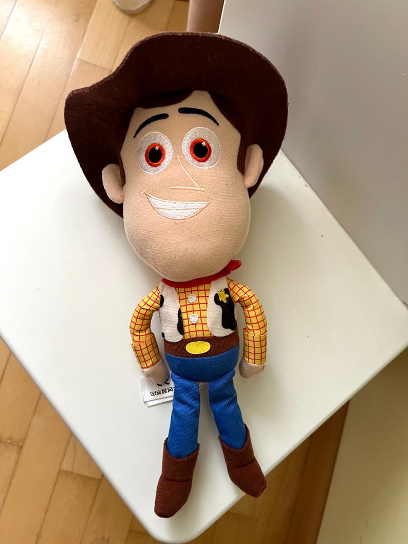 Puede incluir: Un peluche de un personaje de dibujos animados de vaquero. El juguete tiene un sombrero marr&oacute;n, una cara beige con una expresi&oacute;n sonriente y un pa&ntilde;uelo rojo. El personaje viste una camisa a cuadros amarilla, un chaleco blanco, pantalones azules y botas marrones.