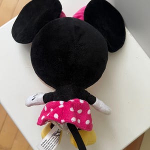 May include: A plush Minnie Mouse doll, viewed from the back. The doll features large black ears, a pink bow, and a pink skirt with white polka dots. The doll has white gloves and yellow shoes. A tag is visible.