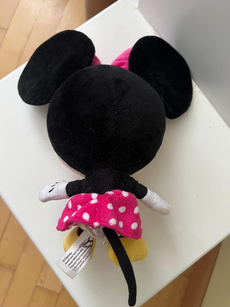 May include: A plush Minnie Mouse doll, viewed from the back. The doll features large black ears, a pink bow, and a pink skirt with white polka dots. The doll has white gloves and yellow shoes. A tag is visible.