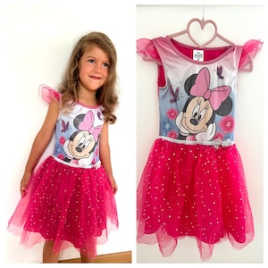 Puede incluir: Un vestido rosa de Minnie Mouse con falda de tul y un estampado de Minnie Mouse en el corpiño. El vestido tiene mangas con volantes y una cinta plateada en la cintura. El vestido está colgado de una percha rosa en forma de corazón.