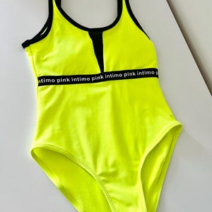 Op de afbeelding: Neon gele badpak met zwarte afwerking en een zwart mesh paneel bij de halslijn. Het badpak heeft de tekst "intimo pink" gedrukt op een band over de borst. Het badpak heeft een heldere, opvallende kleur.