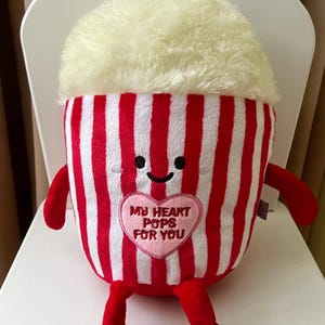 May include: A soft popcorn toy with a red and white striped container, red arms and legs, and a fluffy, cream-coloured top. A pink heart with the text "MY HEART POPS FOR YOU" is on the front. The toy has a smiling face.