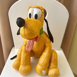 Puede incluir: Un peluche de Pluto, un perro amarillo con orejas y nariz negras, sentado en una silla blanca. El juguete tiene un collar verde, una lengua roja y ojos blancos. Es un personaje clásico de Disney.