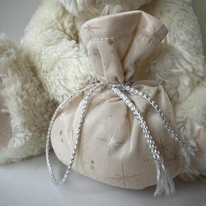 May include: A small, beige fabric gift bag with a silver drawstring closure. The bag is decorated with a subtle star pattern and is tied with a silver cord. The bag is in front of a fluffy, cream-colored teddy bear.