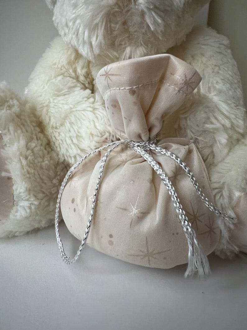 May include: A small, beige fabric gift bag with a silver drawstring closure. The bag is decorated with a subtle star pattern and is tied with a silver cord. The bag is in front of a fluffy, cream-colored teddy bear.