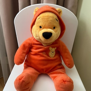 Può includere: Un peluche di Winnie the Pooh, seduto su una sedia bianca. L'orso indossa un completo arancione con cappuccio e una piccola grafica ricamata di Winnie the Pooh sul petto. L'orso ha un naso nero e un'espressione amichevole.