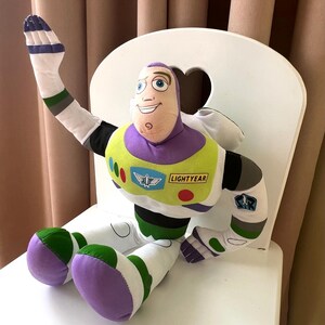 May include: A plush Buzz Lightyear doll, featuring a purple and white space suit with green and yellow accents. The doll has a smiling face and is seated on a white chair. The word "LIGHTYEAR" is visible on the chest.