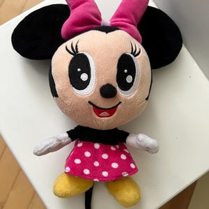 May include: A plush Minnie Mouse doll with a pink bow, black ears, and a pink dress with white polka dots. The doll has yellow shoes and white gloves. The doll is a popular character from Disney.
