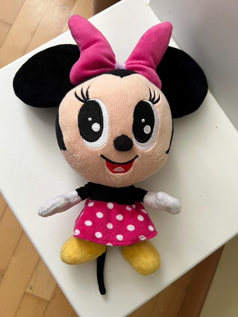 May include: A plush Minnie Mouse doll with a pink bow, black ears, and a pink dress with white polka dots. The doll has yellow shoes and white gloves. The doll is a popular character from Disney.
