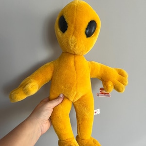 Puede incluir: Un peluche alienígena amarillo con grandes ojos negros ovalados y pequeños brazos y pies. El juguete tiene un lazo en la parte superior para colgarlo. El alienígena se sostiene contra un fondo gris.