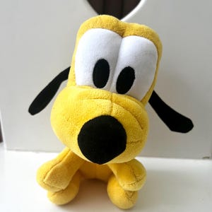 May include: A plush toy of Pluto, a yellow cartoon dog with large white eyes, black pupils, and a black nose. The toy has black floppy ears and is sitting down. The background is a white surface with a heart-shaped cutout.