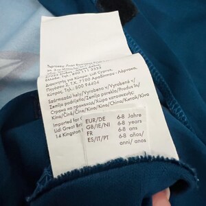 May include: Teal-colored garment with a white tag attached. The tag includes text in multiple languages, indicating the product's origin and size range of 6-8 years. A black velcro patch is visible.