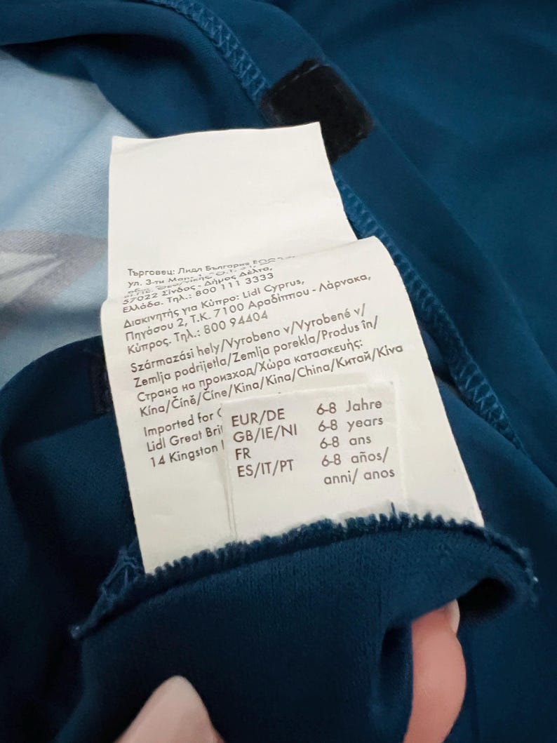 May include: Teal-colored garment with a white tag attached. The tag includes text in multiple languages, indicating the product's origin and size range of 6-8 years. A black velcro patch is visible.