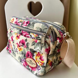 Op de afbeelding: Crossbody tas met bloemenprint op een cr&egrave;mekleurige achtergrond, met roze, gele en paarse bloemen. De tas heeft twee compartimenten met ritssluiting, een beige band en een kleine logopatch.