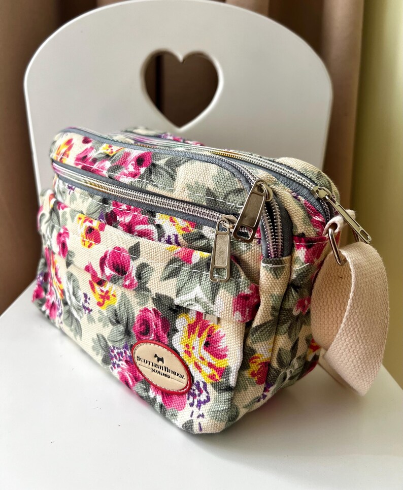 Op de afbeelding: Crossbody tas met bloemenprint op een cr&egrave;mekleurige achtergrond, met roze, gele en paarse bloemen. De tas heeft twee compartimenten met ritssluiting, een beige band en een kleine logopatch.