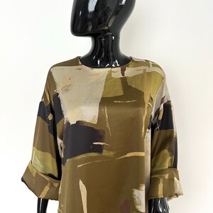 May include: Olive green blouse with a unique abstract pattern in shades of beige, black, and brown. The top features a round neckline and three-quarter sleeves with folded cuffs. The fabric has a silky sheen.