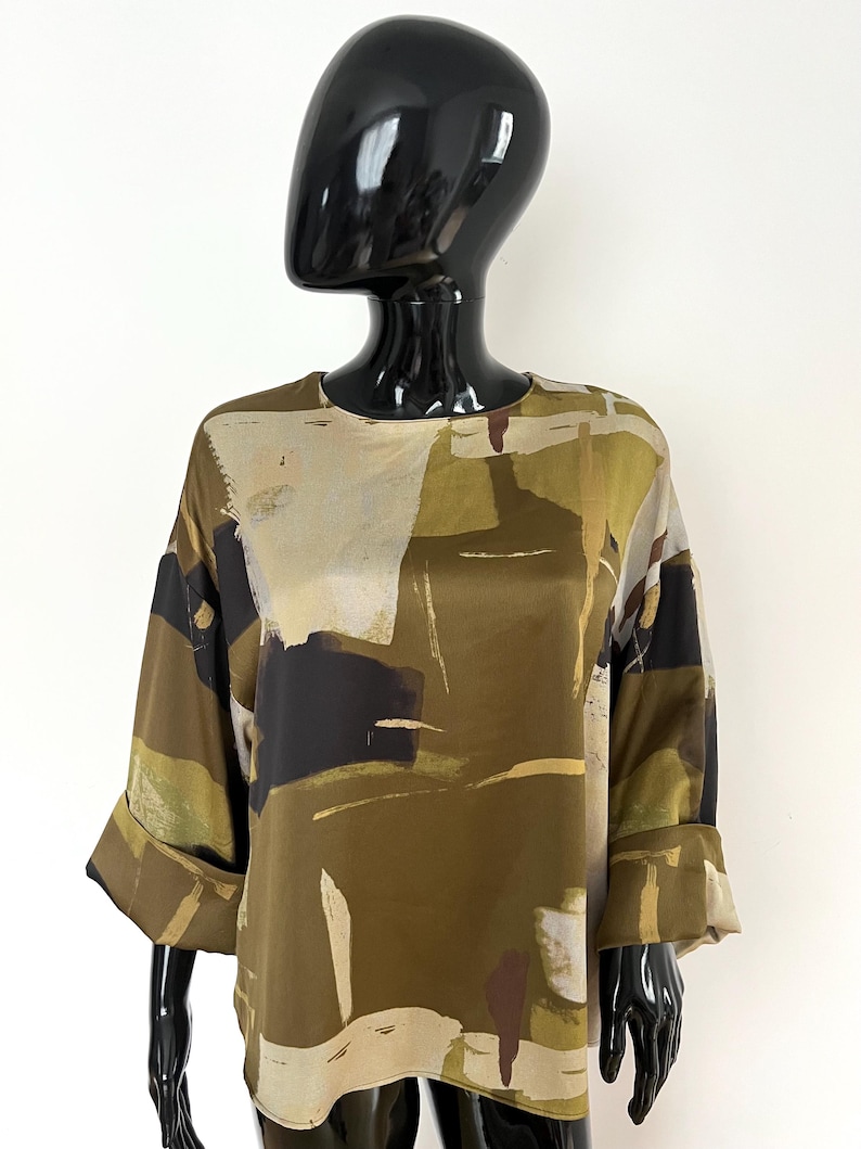 May include: Olive green blouse with a unique abstract pattern in shades of beige, black, and brown. The top features a round neckline and three-quarter sleeves with folded cuffs. The fabric has a silky sheen.