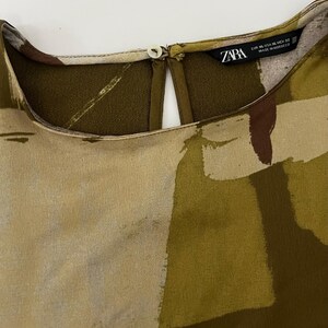 May include: Close-up of a blouse with a keyhole neckline and a button closure. The fabric features an abstract pattern in shades of olive green, beige, and brown. The tag reads "ZARA" with sizing information.