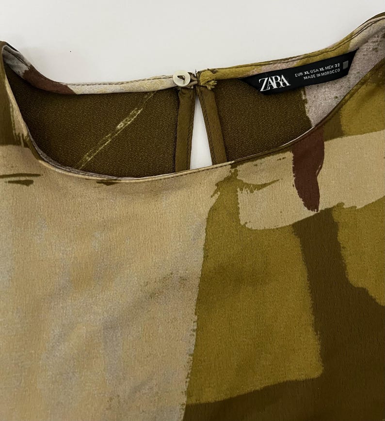 May include: Close-up of a blouse with a keyhole neckline and a button closure. The fabric features an abstract pattern in shades of olive green, beige, and brown. The tag reads "ZARA" with sizing information.