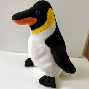 May include: A soft toy penguin with a black head, wings, and feet, a white body, and a yellow and orange beak. The penguin is standing on a white surface.