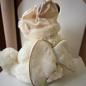 May include: A plush, cream-colored bunny with a drawstring hat and star-patterned wings. The bunny has a fluffy body, a soft hat with a pom-pom, and wings trimmed with gold. The wings are decorated with a subtle star pattern.