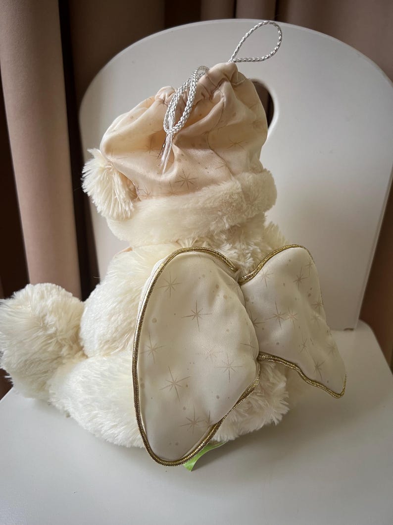 May include: A plush, cream-colored bunny with a drawstring hat and star-patterned wings. The bunny has a fluffy body, a soft hat with a pom-pom, and wings trimmed with gold. The wings are decorated with a subtle star pattern.