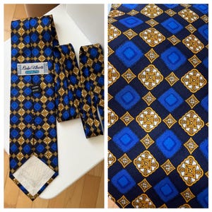 Puede incluir: Una corbata estampada en azul, dorado y negro con un diseño geométrico repetitivo. La etiqueta dice "Lado Alberti, Silk 100%" y está hecha en España. La corbata se muestra sobre una superficie blanca.