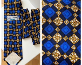 Puede incluir: Una corbata estampada en azul, dorado y negro con un diseño geométrico repetitivo. La etiqueta dice "Lado Alberti, Silk 100%" y está hecha en España. La corbata se muestra sobre una superficie blanca.