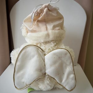 May include: A plush angel doll with cream-colored fur and white wings outlined in gold. The wings are decorated with a star pattern. The doll wears a matching hat with a faux fur trim and a drawstring closure.