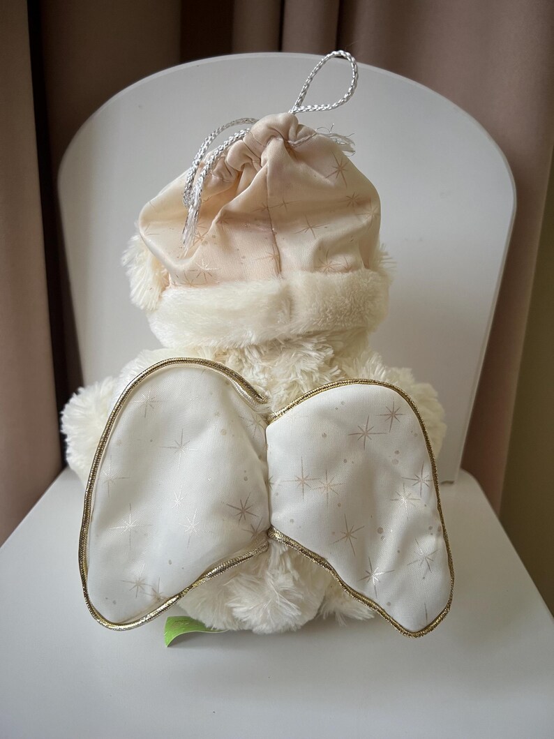 May include: A plush angel doll with cream-colored fur and white wings outlined in gold. The wings are decorated with a star pattern. The doll wears a matching hat with a faux fur trim and a drawstring closure.