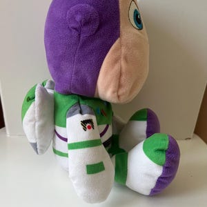 Pode incluir: Um brinquedo de pel&uacute;cia Buzz Lightyear da franquia Toy Story. O brinquedo apresenta uma cabe&ccedil;a roxa, um rosto bege claro e um traje espacial verde e branco. O brinquedo est&aacute; sentado com os bra&ccedil;os e pernas dobrados.