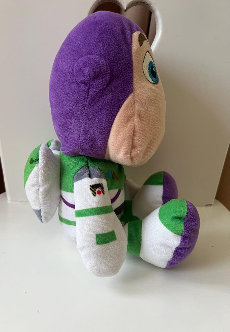 Pode incluir: Um brinquedo de pel&uacute;cia Buzz Lightyear da franquia Toy Story. O brinquedo apresenta uma cabe&ccedil;a roxa, um rosto bege claro e um traje espacial verde e branco. O brinquedo est&aacute; sentado com os bra&ccedil;os e pernas dobrados.