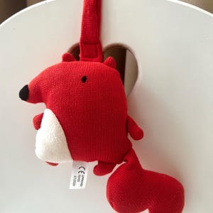 May include: A red knitted fox toy with a white belly and black nose. The fox has a loop for hanging and a rounded tail. The toy is hanging on a white surface.