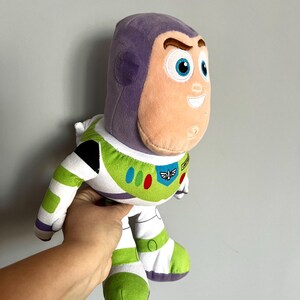 May include: A plush Buzz Lightyear toy from the Toy Story franchise. The toy features a purple helmet, a peach-colored face, and a white and green spacesuit. The toy has blue eyes and a friendly expression.
