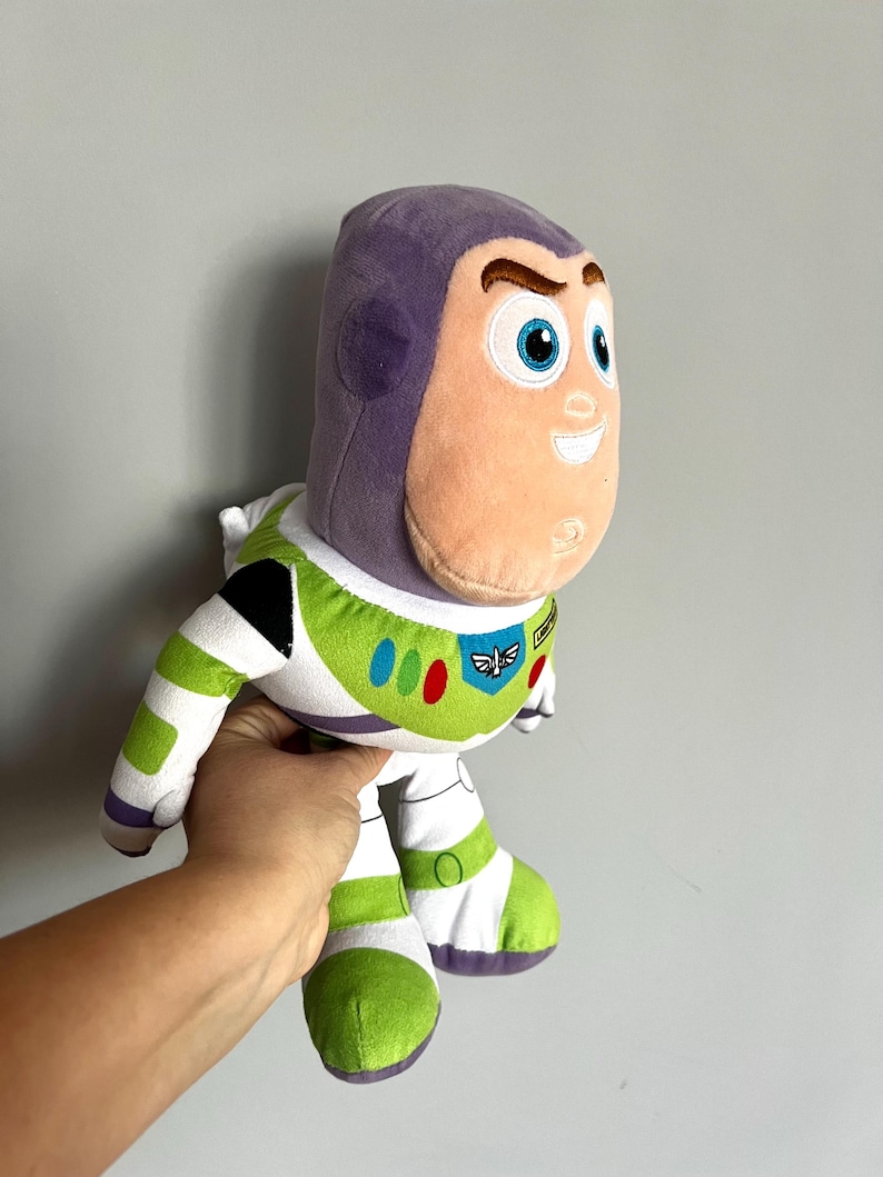 May include: A plush Buzz Lightyear toy from the Toy Story franchise. The toy features a purple helmet, a peach-colored face, and a white and green spacesuit. The toy has blue eyes and a friendly expression.